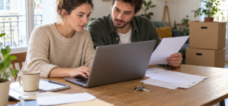 Un couple consulte un ordinateur et des documents à une table, avec des cartons de déménagement en arrière-plan.