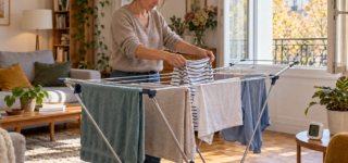 Une femme étend des vêtements sur un séchoir dans un salon, près d’une fenêtre ouverte.