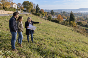 Un couple et une professionnelle avec un plan observent un terrain herbeux en pente, en contrebas d’une route.