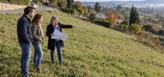 Un couple et une professionnelle avec un plan observent un terrain herbeux en pente, en contrebas d’une route.