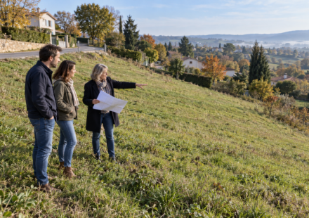 Un couple et une professionnelle avec un plan observent un terrain herbeux en pente, en contrebas d’une route.