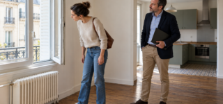 Une candidate à la location examine une fenêtre ouverte pendant la visite d’un appartement avec un professionnel immobilier.